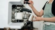 Crop faceless young female barista in apron steaming milk at modern coffee machine while preparing delicious latte