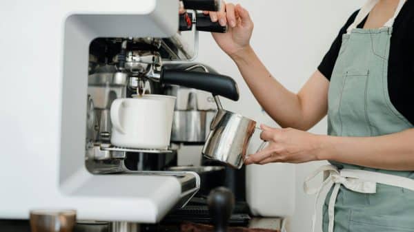 Crop faceless young female barista in apron steaming milk at modern coffee machine while preparing delicious latte