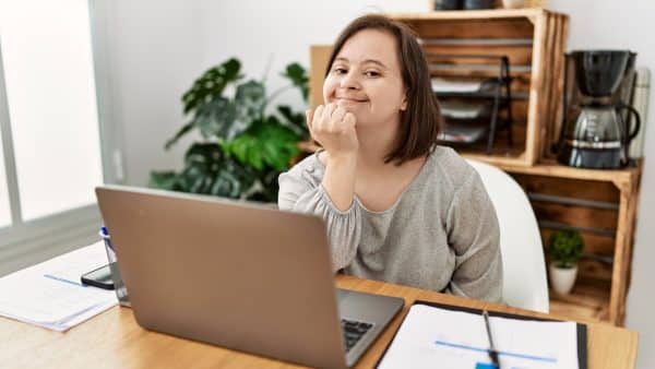 A photo of a woman with a learning disability working at a laptop. She is smiling at the camera.