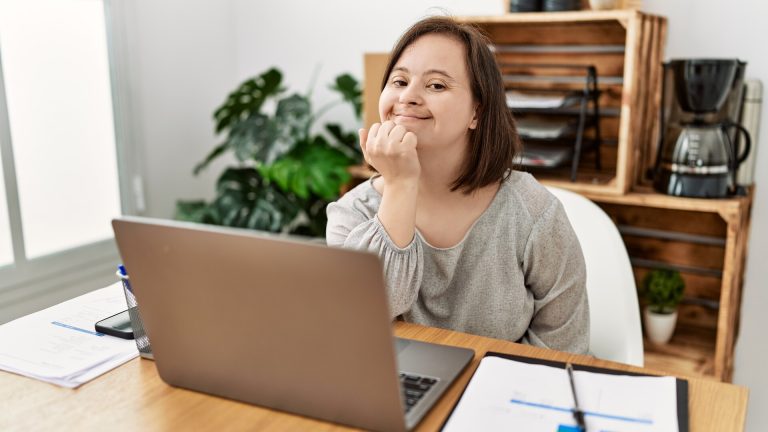 A photo of a woman with a learning disability working at a laptop. She is smiling at the camera.