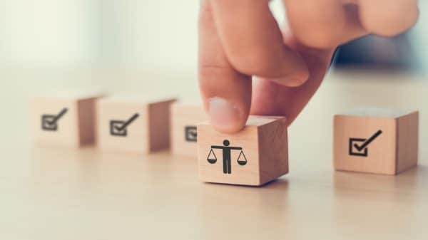 A hand picking up a wooden block with a person holding scales icon, placed among other blocks with checkmark icons, symbolising fairness or justice in decision-making.